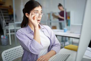 Businesswoman in eyeglasses sitting at the table and looking at monitor with surprise
