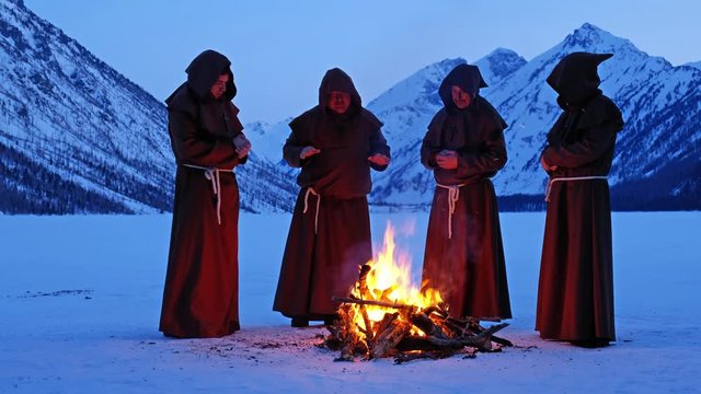 Group of monks in brown cloaks of the order of St. Francis are warming themselves by the fire