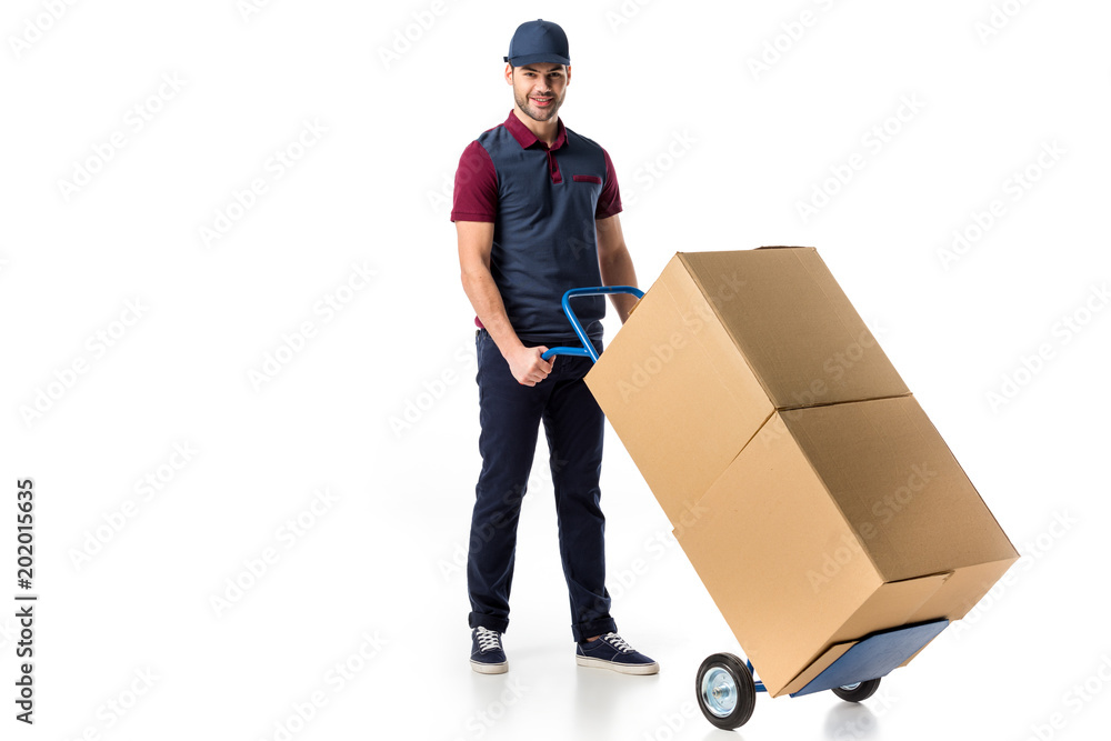 Wall mural smiling delivery man in uniform pushing hand truck with cardboard boxes isolated on white