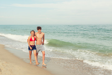 Two teenagers: a girl and a boy with blond hair, dressed in a swimsuit walks returning to the viewer on the sea beach.