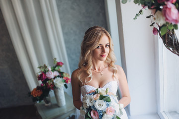 Portrait of a beautiful bride with curly hair in a gray studio with flowers. A sweet bride stands near a window in a modern studio.