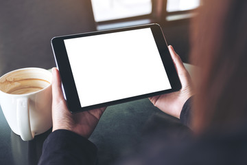 Mockup image of hands holding black tablet pc with blank white screen and coffee cup on table in...