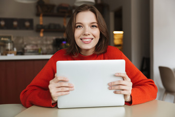 Photo of content young woman 20s in sweater looking at camera and holding silver laptop in hands, while sitting in restaurant