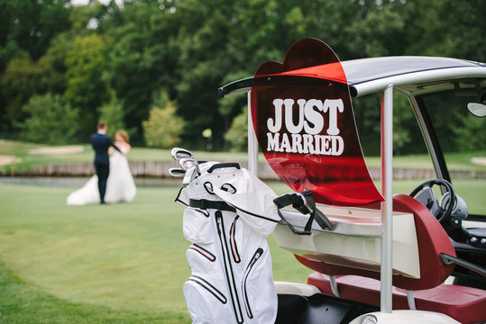 Just married sign on golf cart with dancing wedding couple in blur on background, copy space. Car for golf with equipment and bride and groom on golf course
