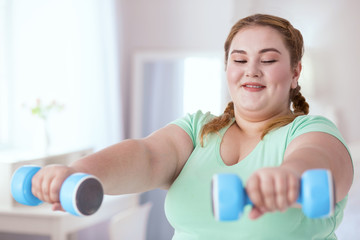 Sports program. Smiling young woman doing exercises with dumbbells while watching sports program