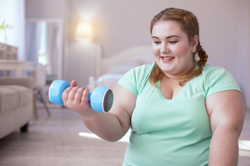 Muscles gain. Plump young woman holding dumbbell while sitting on the yoga mat