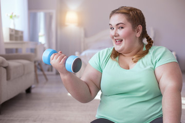 Dumbbells exercises. Fascinated plump woman sitting on the yoga mat while doing exercises