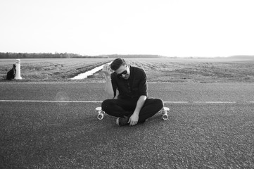 A young man in sunglasses smiling and sitting on a longboard,