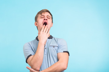 Sleepy young man yawning, waking up closing his mouth with hand isolated on blue background