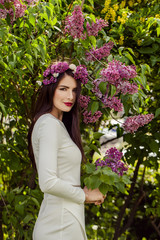 Beautiful fashion model in white dress outdoors. Perfect young woman with lilac flowers in spring park