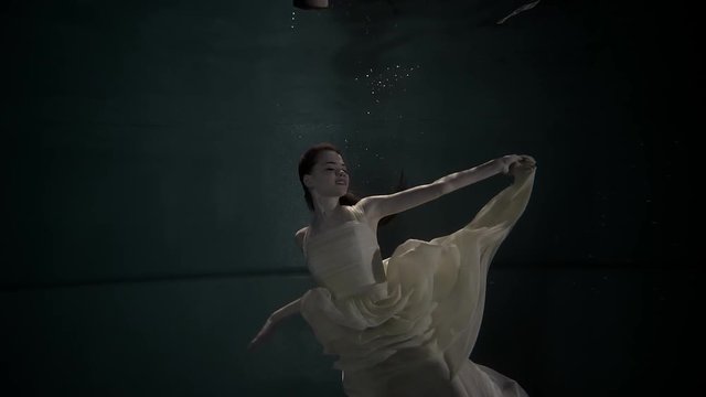 Beautiful young girl posing underwater alone in a white dress. Happy girl floating in darkness. Charming model