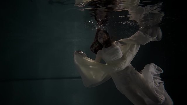 Beautiful red haired woman floating in darkness under the water. She is wearing white dress. Reflection on the surface.