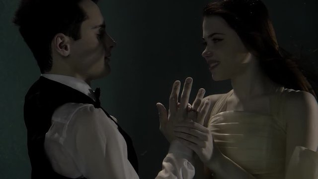 Portrait of a lovers marrying underwater in darkness. Wedding dress on a woman and suit on a man. Putting ring on his finger.