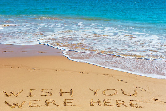 WISH YOU WERE HERE insctiption on wet beach sand with the turquoise sea on background