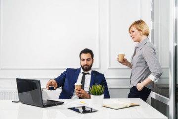 Successful business colleagues holding cups of coffee work with laptop in white office interior