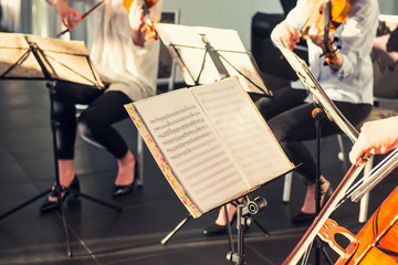 Selective Focus on Music note sheets on stand with background of playing cellists and violinists band on event in bright day sunlight. Commercial music. Service at business meeting, party, weddings.