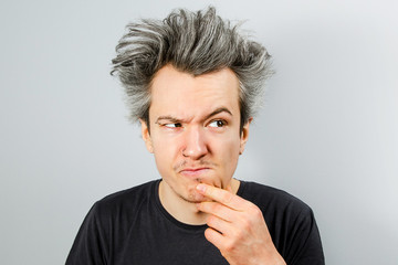 Unshorn and unshaven young guy with piercings on his face, thinking, holding hand at the face and looking aside, on gray background.