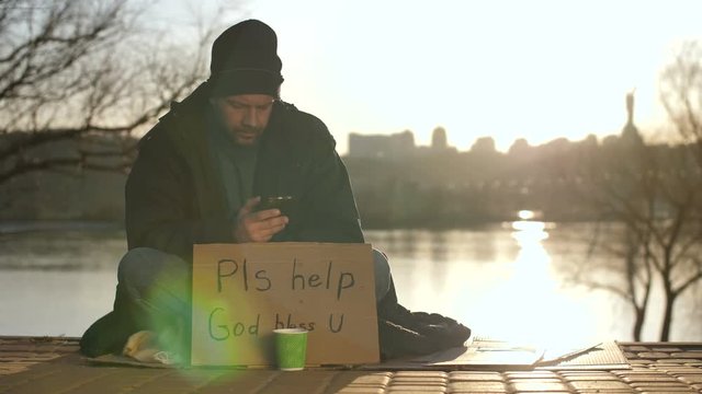 Mature homeless bearded man in hat and jacket sitting on street during sunset with cardboard sign and paper cup, and using his mobile phone at the river bank. Social issues concept, dolly shot.