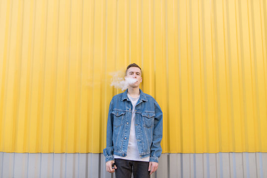 A young man smokes an electronic cigarette on a yellow background. The steam from the wipes comes out of the mouth of a young man