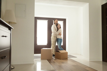 Happy young millennial couple hugging standing near boxes moving into new home, smiling homeowners embracing in modern hallway just moved in own house, mortgage investment and relocation concept