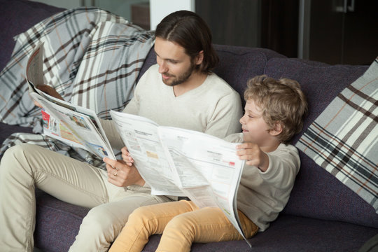 Cute kid son holding newspaper sitting on sofa with dad, curious funny clever boy pretending reading news copying father on weekend, happy daddy enjoying spending time having fun with child at home