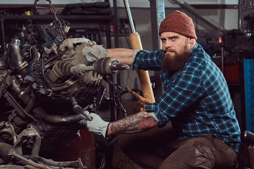 A brutal tattooed bearded mechanic specialist repairs the car engine which is raised on the hydraulic lift in the garage. Service station.