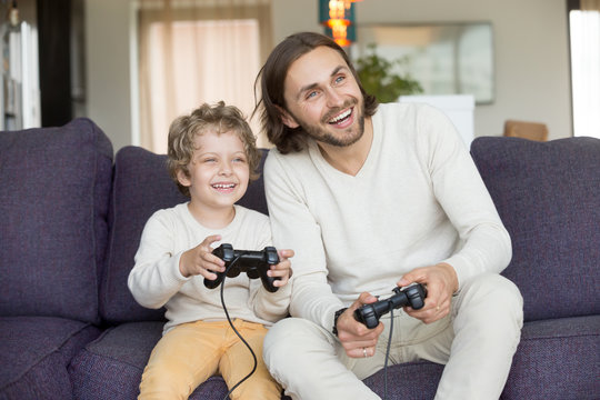 Happy dad laughing playing video game with excited son at home, smiling father having fun with kid boy holding joysticks, child gamer enjoying videogame spending time with daddy together on weekend