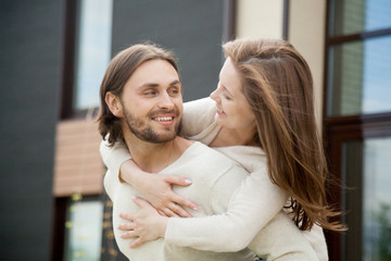 Young millennial couple embracing outdoor, smiling family spending time outside house having fun looking at each other with love affection, caring husband piggybacking wife enjoying leisure together