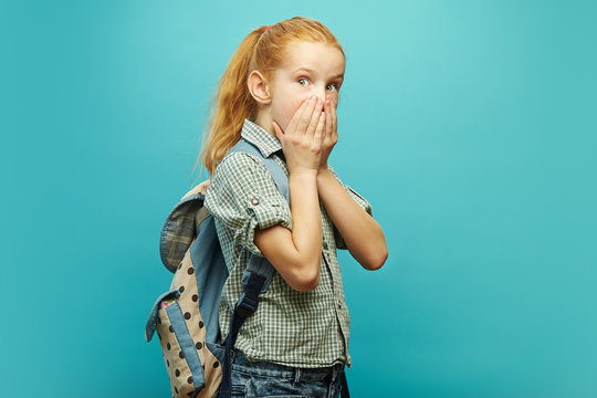 Portrait of emotional girl in uniform and backpack on shoulders, covered her mouth with hands, expresses surprise and fear, shows big eyes, on isolated blue background.