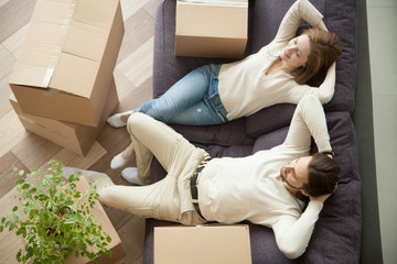Relaxed smiling couple resting on couch moved in new home with boxes, happy real estate renters owners relaxing on sofa in living room, easy relocation with moving delivery service concept, top view