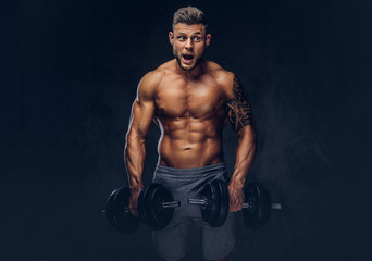 Obraz na płótnie Canvas A handsome shirtless tattooed bodybuilder with stylish haircut and beard, wearing sports shorts, posing in a studio. Isolated on a dark background
