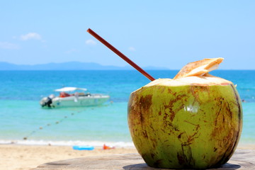 Young green coconut with drinking brown straw on the beach against the azure sea background with blurred white boat.