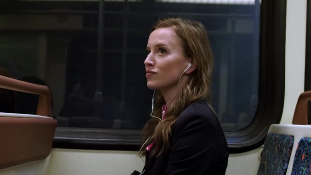 Medium close up, smartly dressed business woman sitting on subway train, listening to audio on small headphones. Hand-held, slow motion HD 60fps.