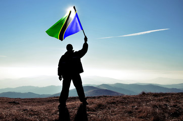 Successful silhouette man winner waving Tanzania flag on top of the mountain peak