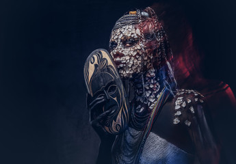 Make-up concept. Portrait of a scary African shaman female with a petrified cracked skin and dreadlocks, holds a traditional mask on a dark background. Make-up concept.