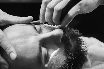 Bearded male sitting in an armchair in a barber shop while hairdresser shaves his beard with a dangerous razor.