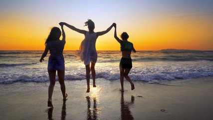 Fototapeta premium Group of people running on the beach in sunset holding for hands having fun jumping and spraying, cinematic steadicamshot