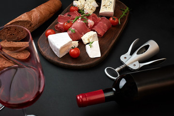 Obraz na płótnie Canvas Red wine, cheese, cherry tomato, bread and prosciutto on wooden board over black backdrop, top view, copy space