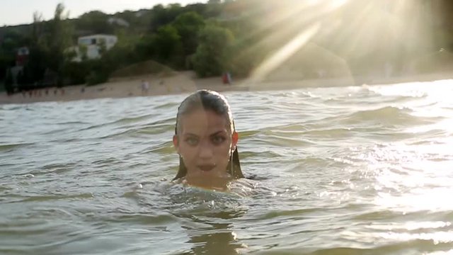 Attractive woman with big breasts emerges from water in a sea water on sunset. Pretty wet smiling girl wearing swimsuit and looks to the camera. Slow motion.