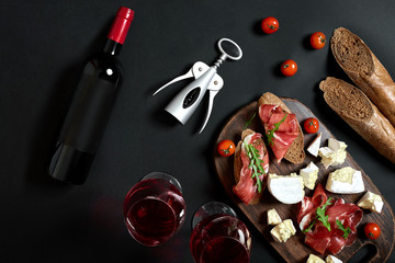 Red wine, cheese, cherry tomato, bread and prosciutto on wooden board over black backdrop, top...