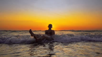 Young smiling woman sitting in sea waves and relaxing at summer vacation at sunset