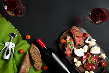 Obraz na płótnie Canvas Prosciutto, salami, baguette slices, tomatoes and nutson rustic wooden board, two glasses of red wine over black background