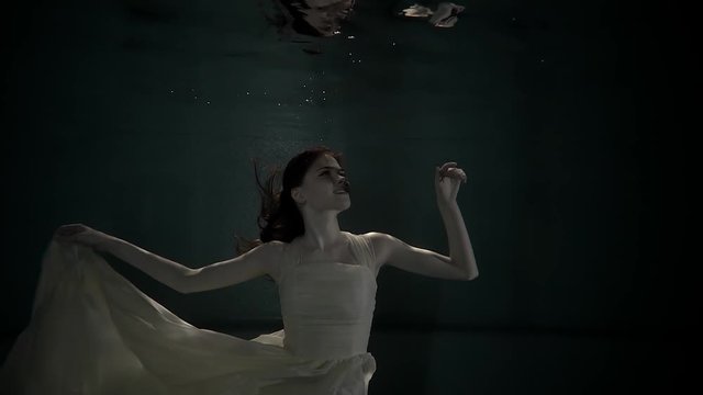 Charming ginger woman is swimming in dark underwater space of aquarium. She is playing with her long dress and her hair is swaying around head