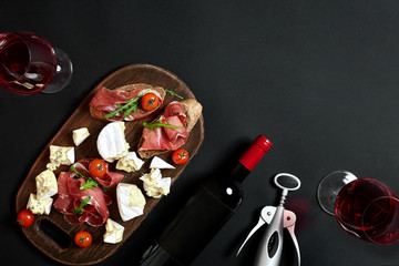Obraz na płótnie Canvas Prosciutto, salami, baguette slices, tomatoes and nutson rustic wooden board, two glasses of red wine over black background