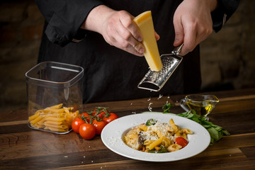 Male chef  hands close-up rubbed Parmesan cheese on a ready penne pasta in a white plate, on a wooden table with cherry tomatoes, basil and olive oil. - 202106085