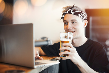 Young smiling man holding glass of beer and working on laptop.