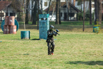 Boy in the camouflage holds a paintball gun  in one hand and protective helmet , standing on the field with group of players on the background