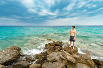 Attractive young shirtless athletic man lost on isolated island standing on rock by water on ocean or sea shore, wearing shorts, looking at sea horizon, travel concept