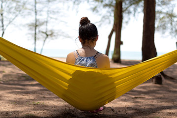 Young teenager girl with long hair but tie it up is sitting on yellow hammock facing with the sea on her relaxing holiday travel