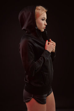 Beautiful seductive sporty girl with the slim sexy figure in the black hoodie, sport underwear and little black shorts is posing in the studio